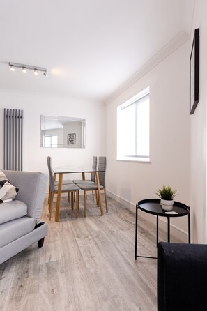 A modern living room with a grey sofa, a dining table with chairs, a small black side table with a plant, and a large window providing natural light.