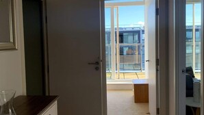 Une pièce avec un plancher en bois, une porte blanche donnant sur un balcon et une vue sur un bâtiment en arrière-plan.