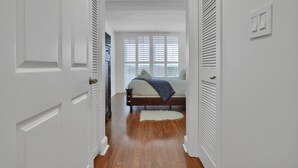 A narrow hallway leading to a bedroom with a bed, a window with blinds, and a white door. - SF 1Bed 1Bath Steps to Marina & CocoWalk in Grove (Miami)