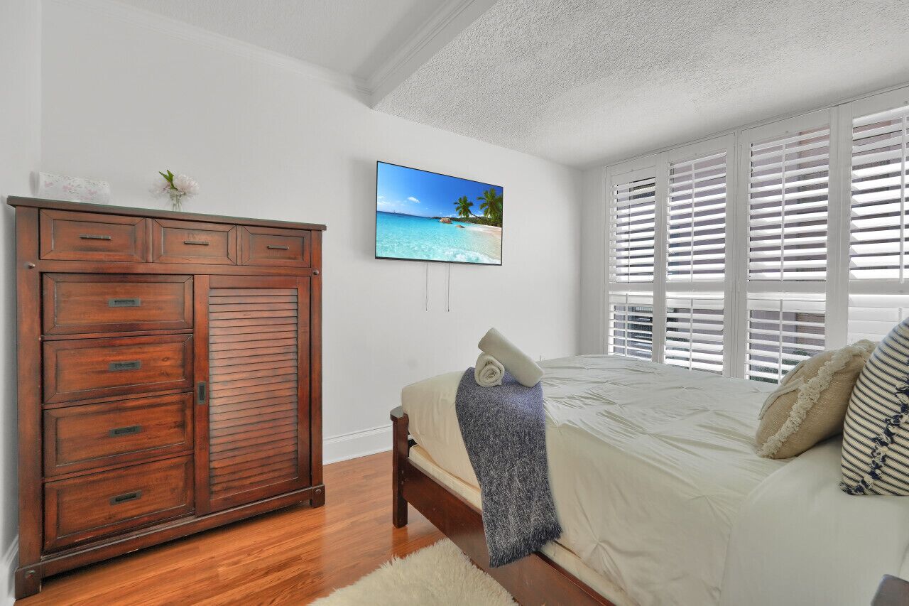 Un dormitorio con un armario de madera, una cama con colcha blanca, un adorno de temática playera y una ventana con persianas blancas.