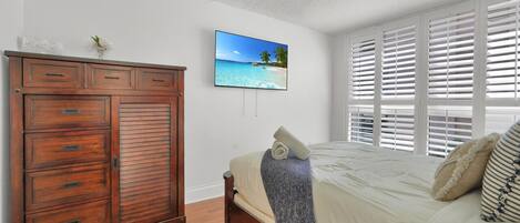 Un dormitorio con un armario de madera, una cama con colcha blanca, un adorno de temática playera y una ventana con persianas blancas.