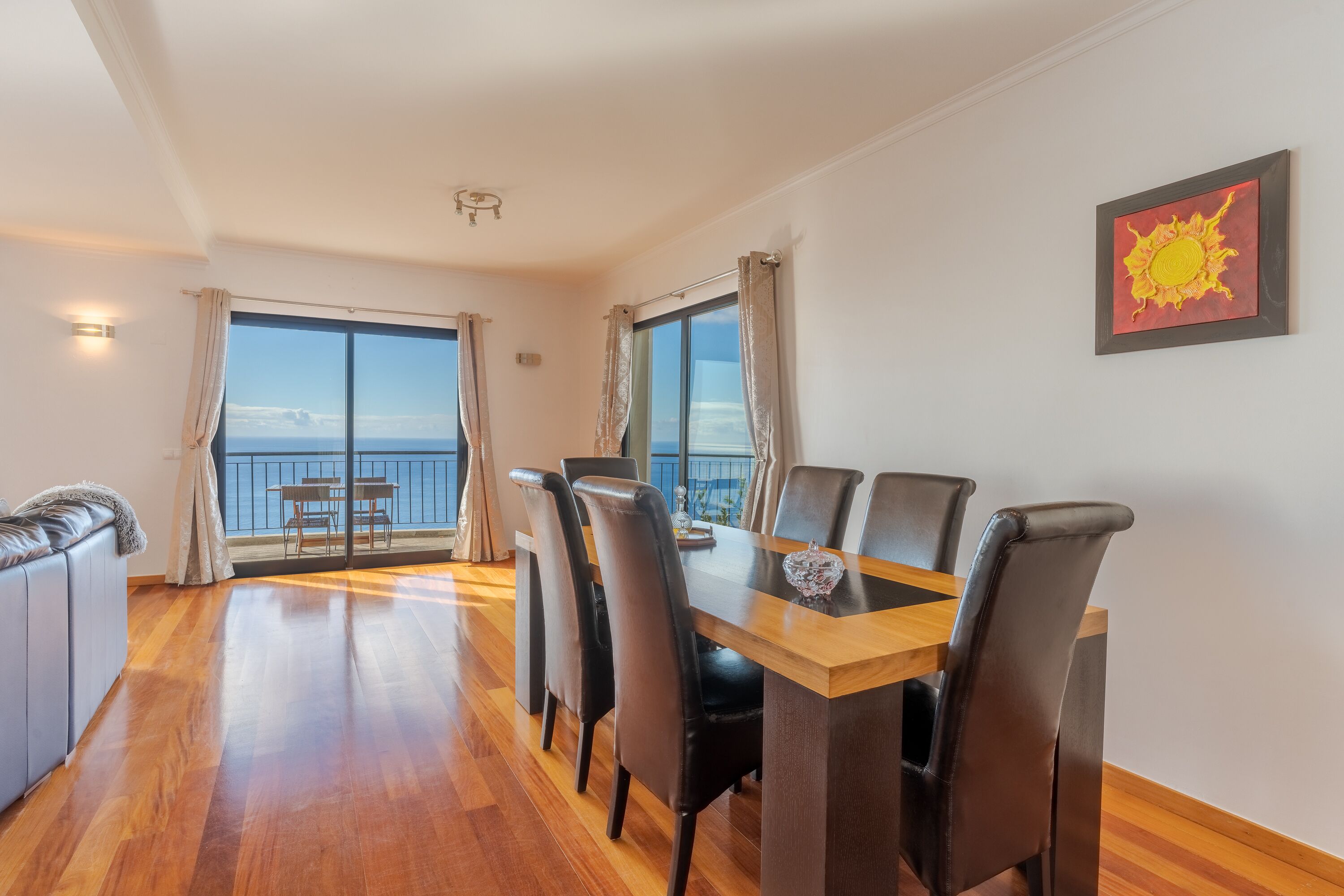 A dining area with a wooden table and chairs, large windows with curtains, and a view of the sea.