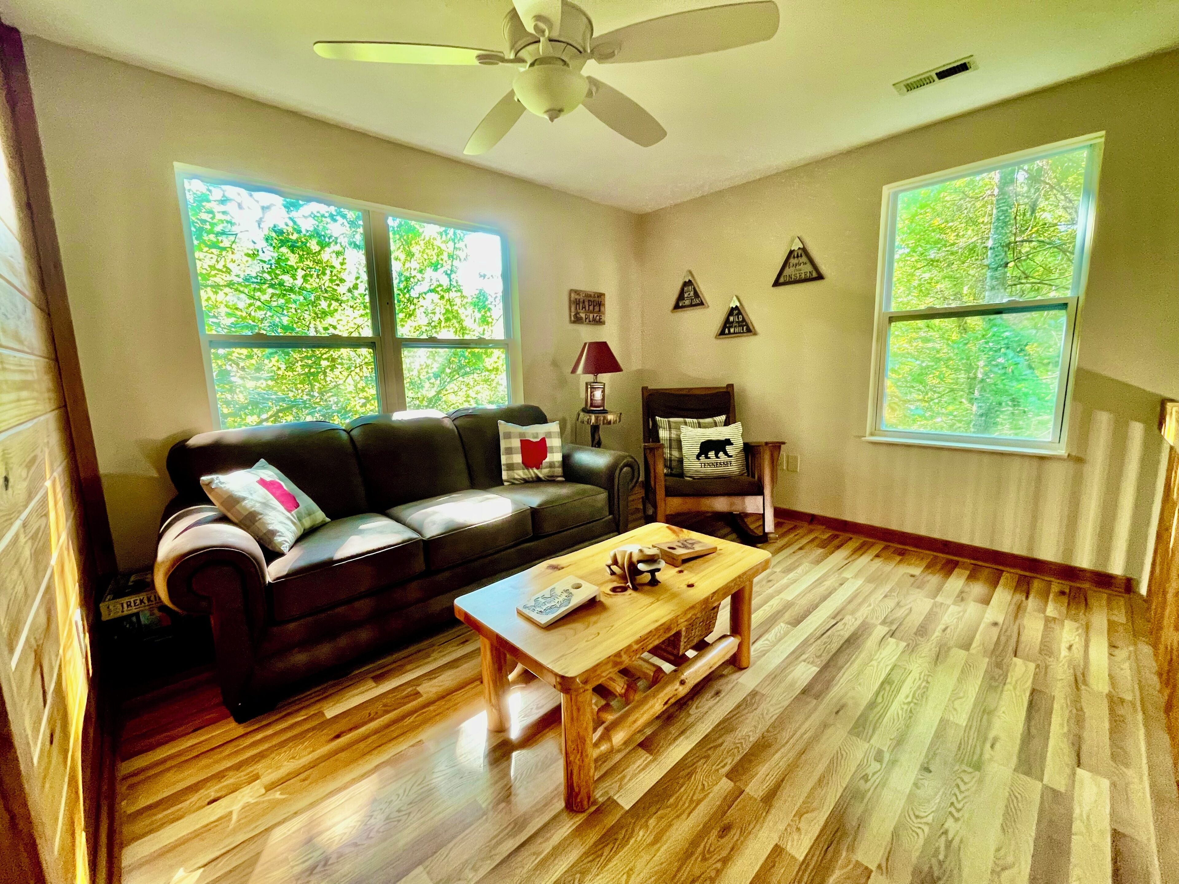 A living room with a dark leather sofa, a wooden coffee table, and a ceiling fan. There are two windows with a view of trees outside.