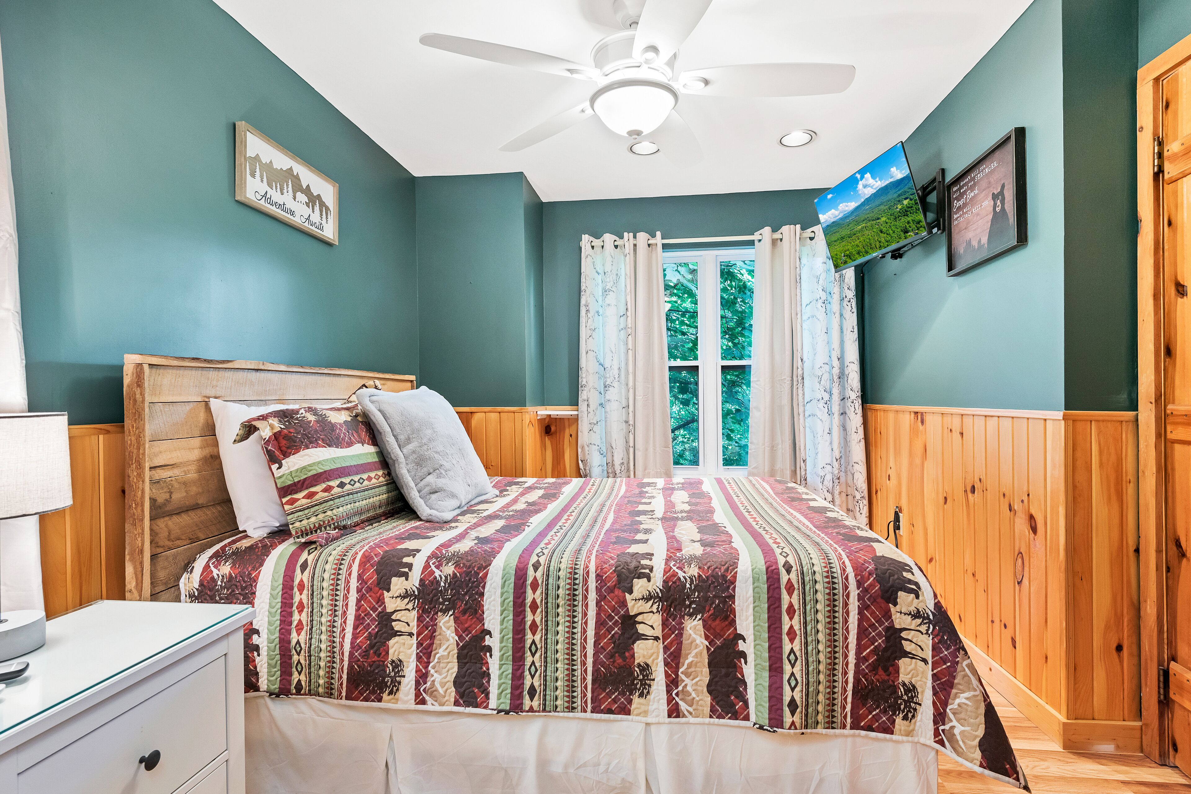 A bedroom with a bed, wooden headboard, a ceiling fan, a window with curtains, and a wall-mounted TV.