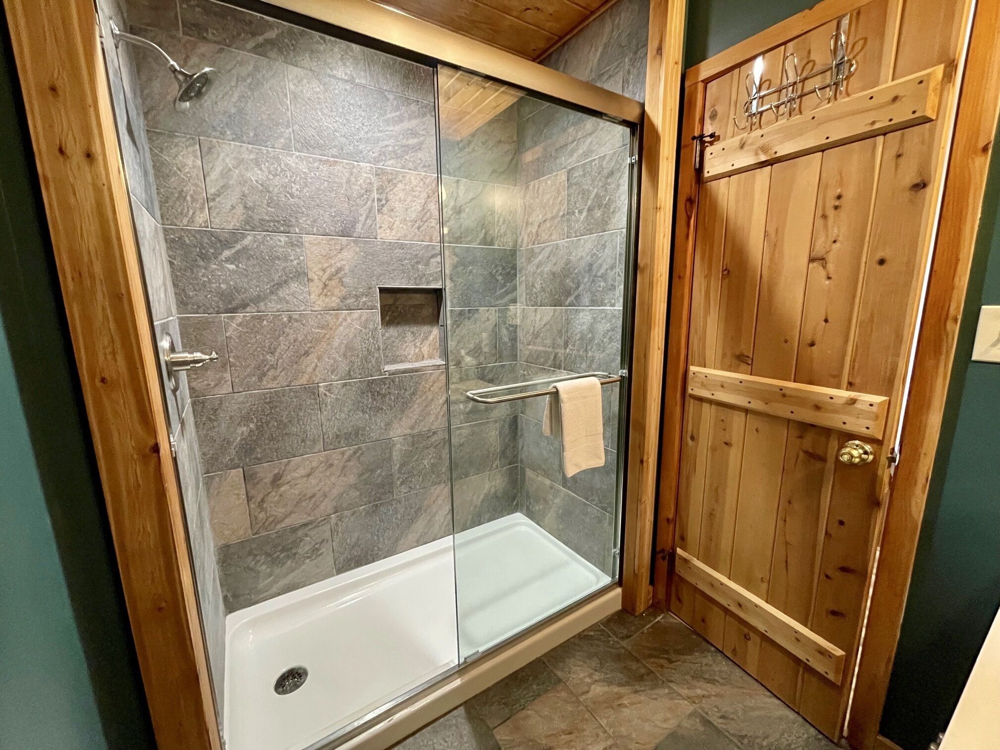 Un espace douche clos par des parois de verre, avec une baignoire, une porte en bois dotée de crochets et des carreaux de pierre.