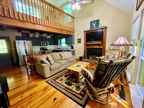 A spacious living room with a high ceiling, a wooden ceiling fan, a fireplace, a sofa, a coffee table, and a wooden chair.