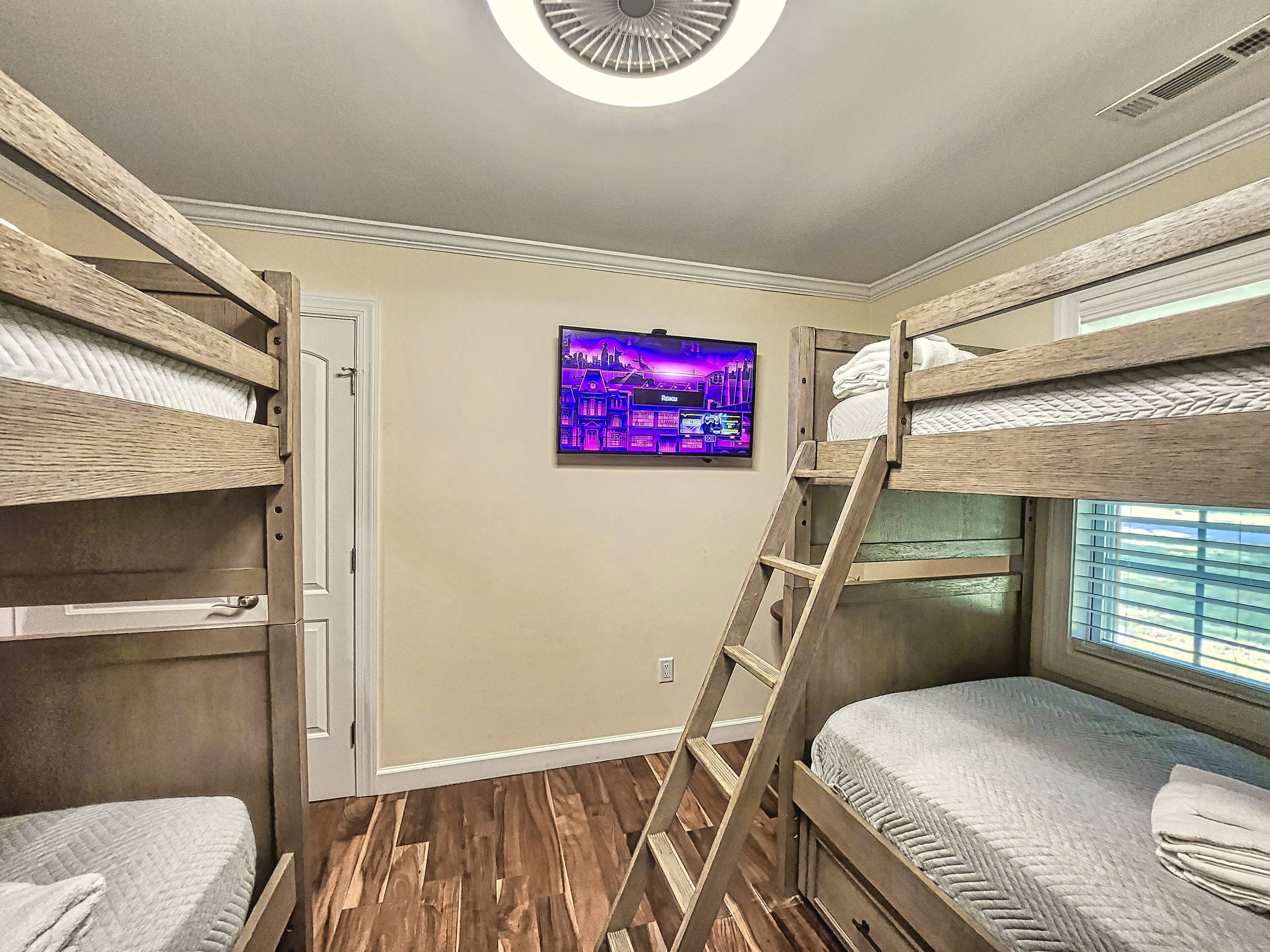 A bunk bed room with a TV on the wall, wooden flooring, and a window with blinds.