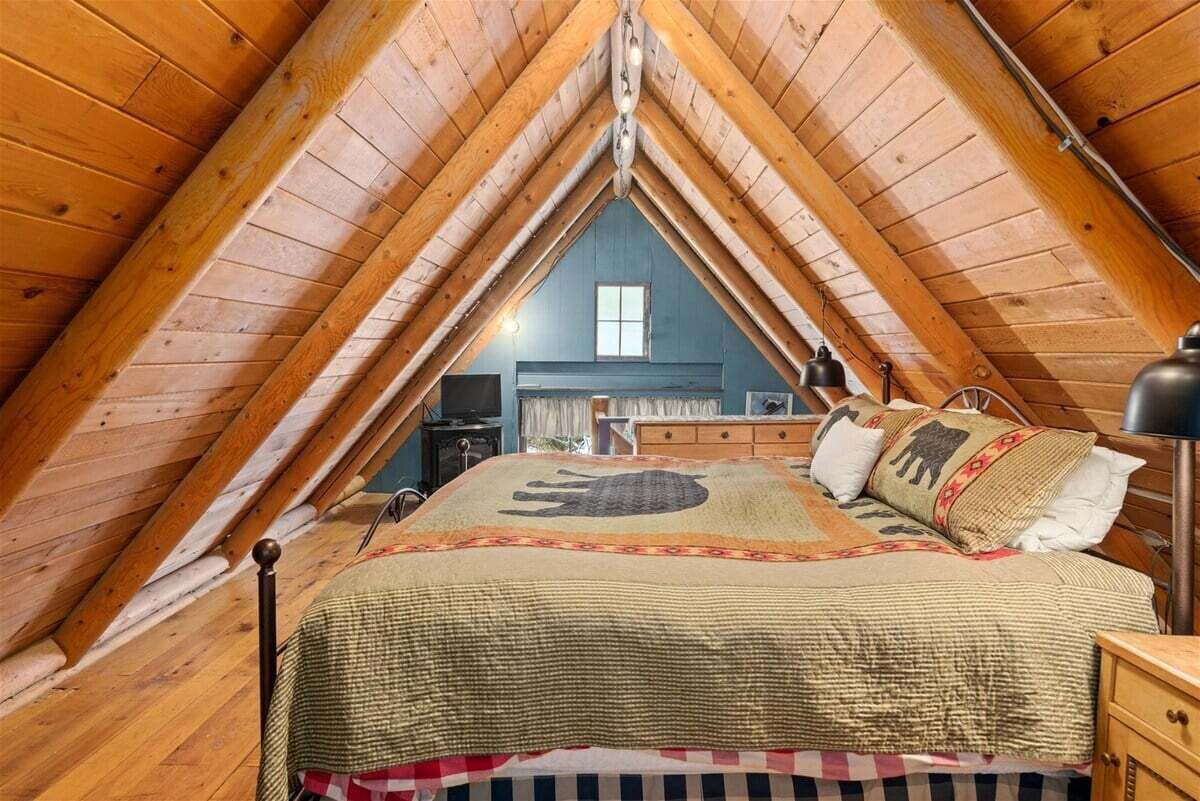 Une chambre mansardée chaleureuse, avec un plafond en bois incliné, un lit avec une literie à motifs, une commode et un petit bureau avec une lampe.