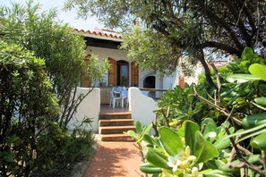 A pathway leading to a building with a white facade and wooden shutters, a small outdoor seating area, and lush greenery.