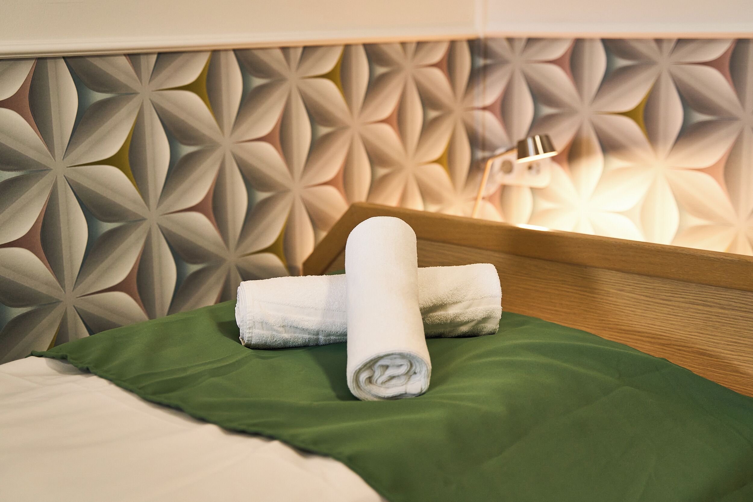 A neatly made bed with a green bedspread and a white towel rolled up at the head.