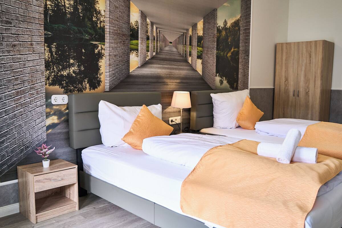 A hotel room with a bed, bedside table, lamp, and a mural of a forest path.