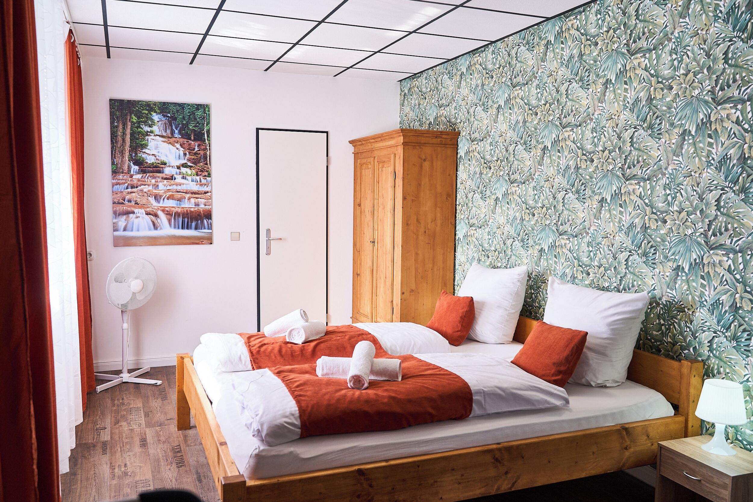 Een hotelkamer met een bed, een houten kledingkast, een ventilator en een muur met behang met bladeren.