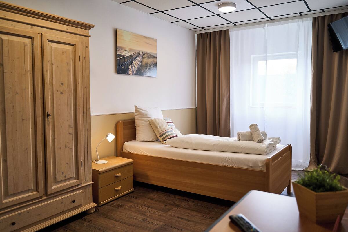 A hotel room with a wooden wardrobe, a bed with white linens, a bedside table with a lamp, and a window with curtains.