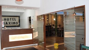 A hotel reception area with a modern design, including a reception desk, a sign that reads "HOTEL MAXIMO", and a glimpse of the interior dining area with tables and chairs. - Hotel Maximo (Pinneberg)