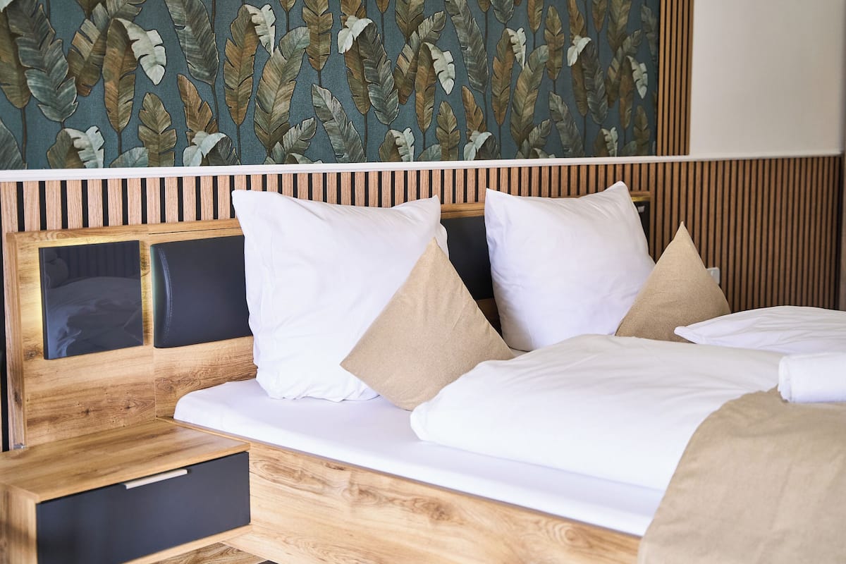 A modern hotel room with a wooden headboard, a large bed with white bedding, and a leaf-patterned wall.