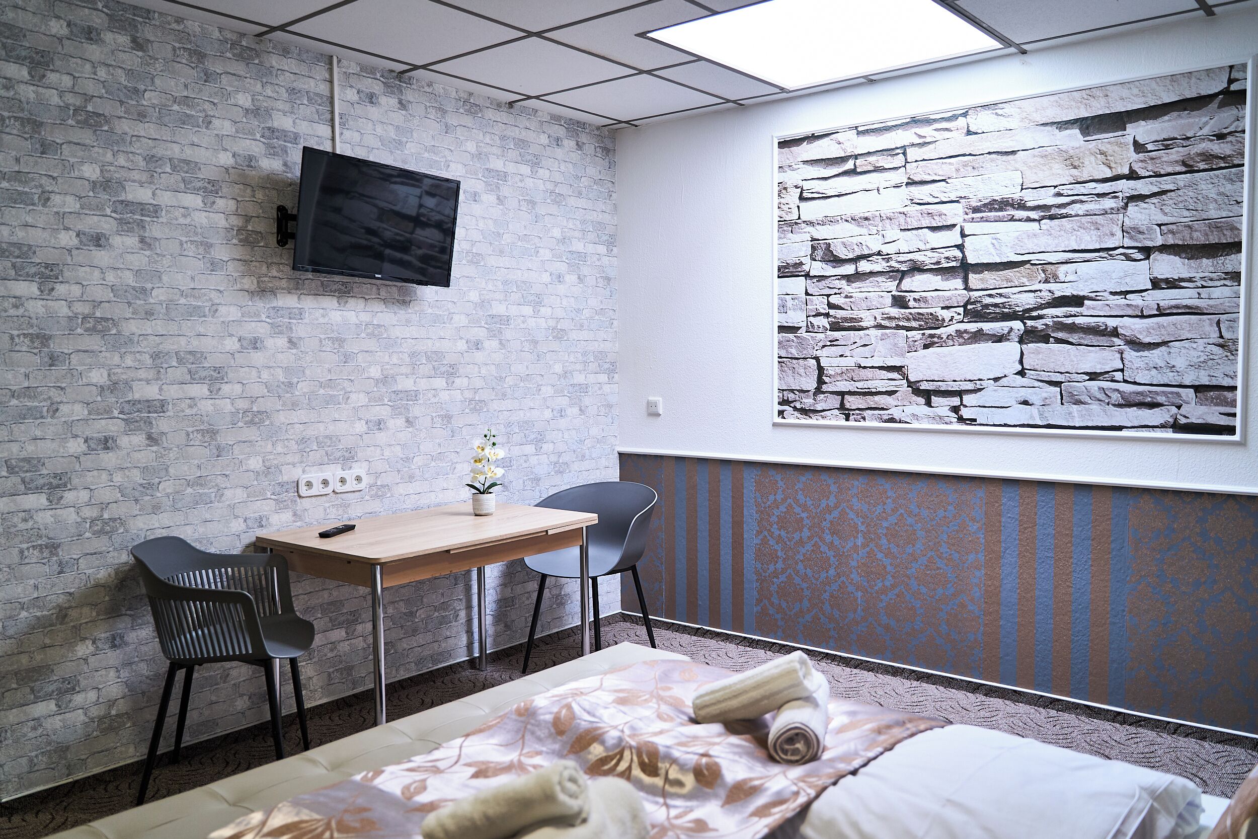 Habitación de hotel con pared de ladrillo, televisor de pantalla plana sobre un escritorio de madera, dos sillas, cama con colcha estampada y un mural de pared de piedra.