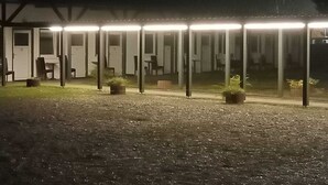 A row of single-room cabins with individual lighting, a gravel ground, and a building in the background. - Hotel Maximo (Pinneberg)