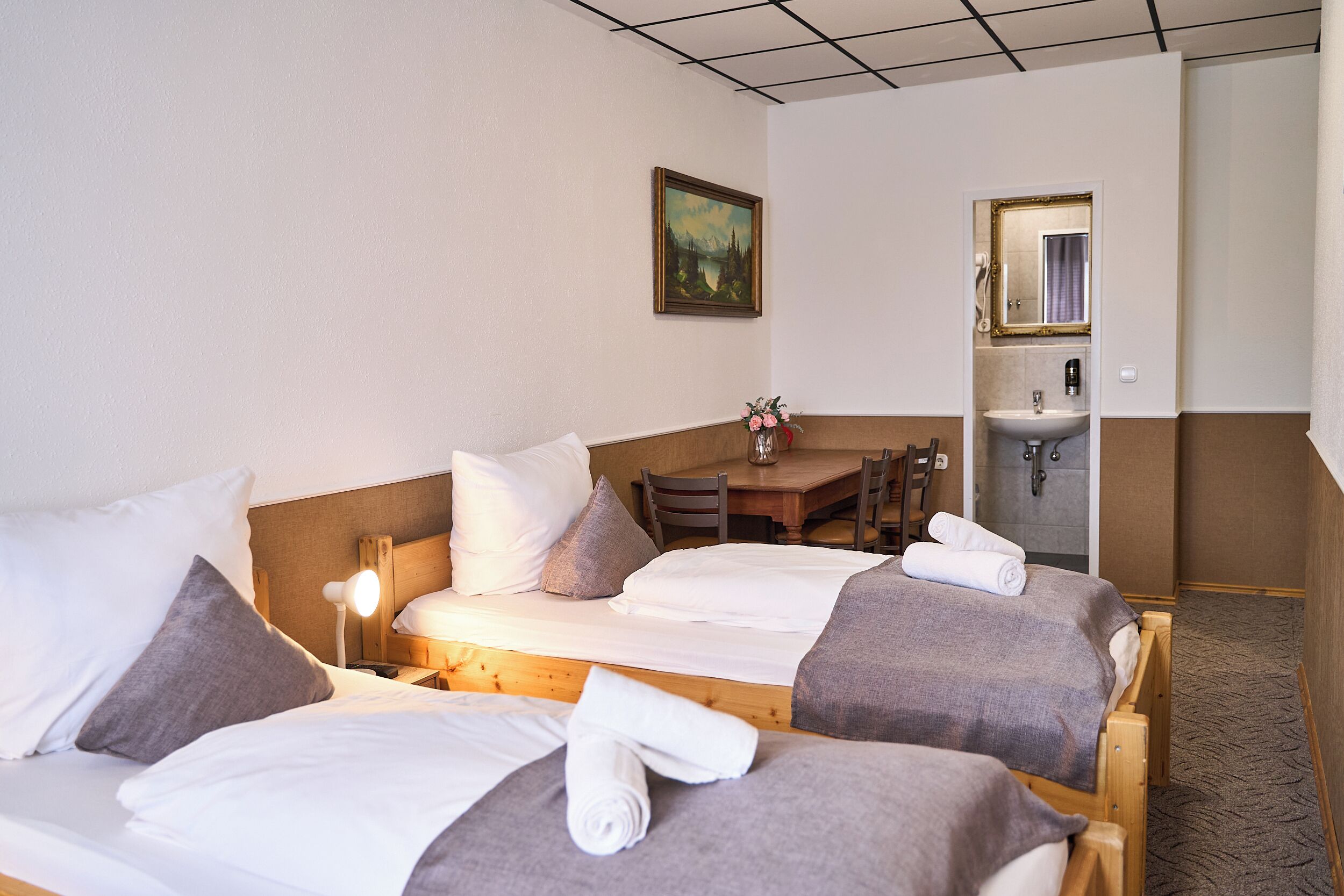 A hotel room with two beds, a small table, a chair, a painting on the wall, and a bathroom with a sink and mirror.