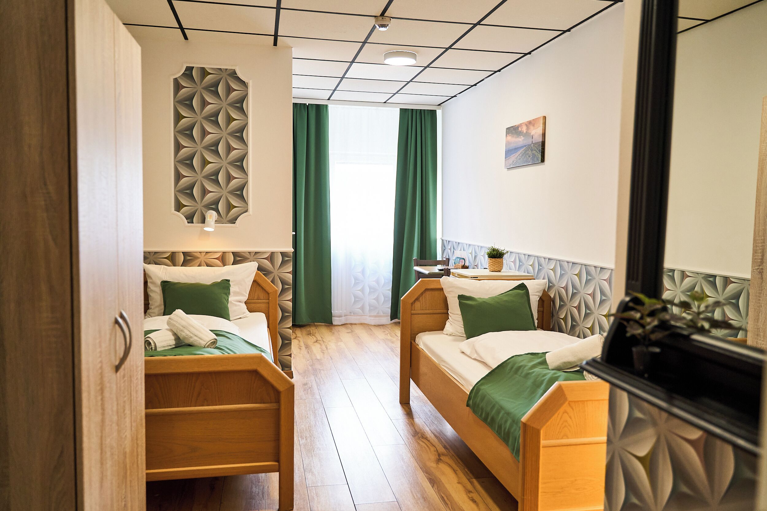 Habitación de hotel con dos camas de madera, una cortina verde, un panel de pared geométrico y una mesita con una planta.