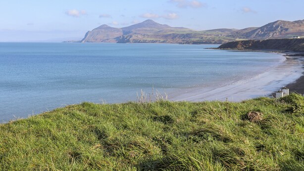 A coastal landscape with a grassy foreground, a sandy beach, and mountains in the background. - Bwthyn Cerrig Man (Pwllheli)