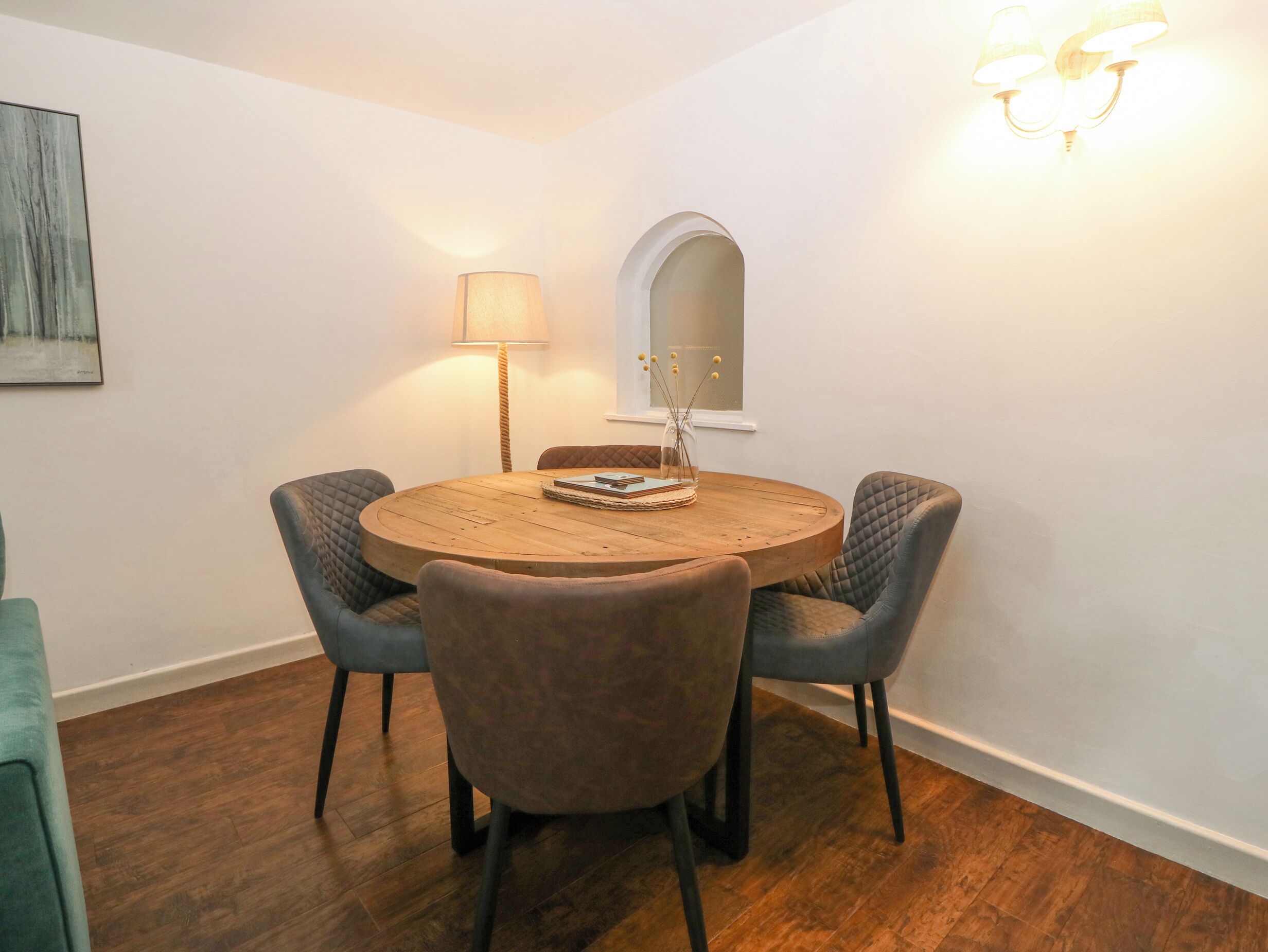 A round wooden table with four cushioned chairs, a floor lamp, and a vase with sticks on the table.
