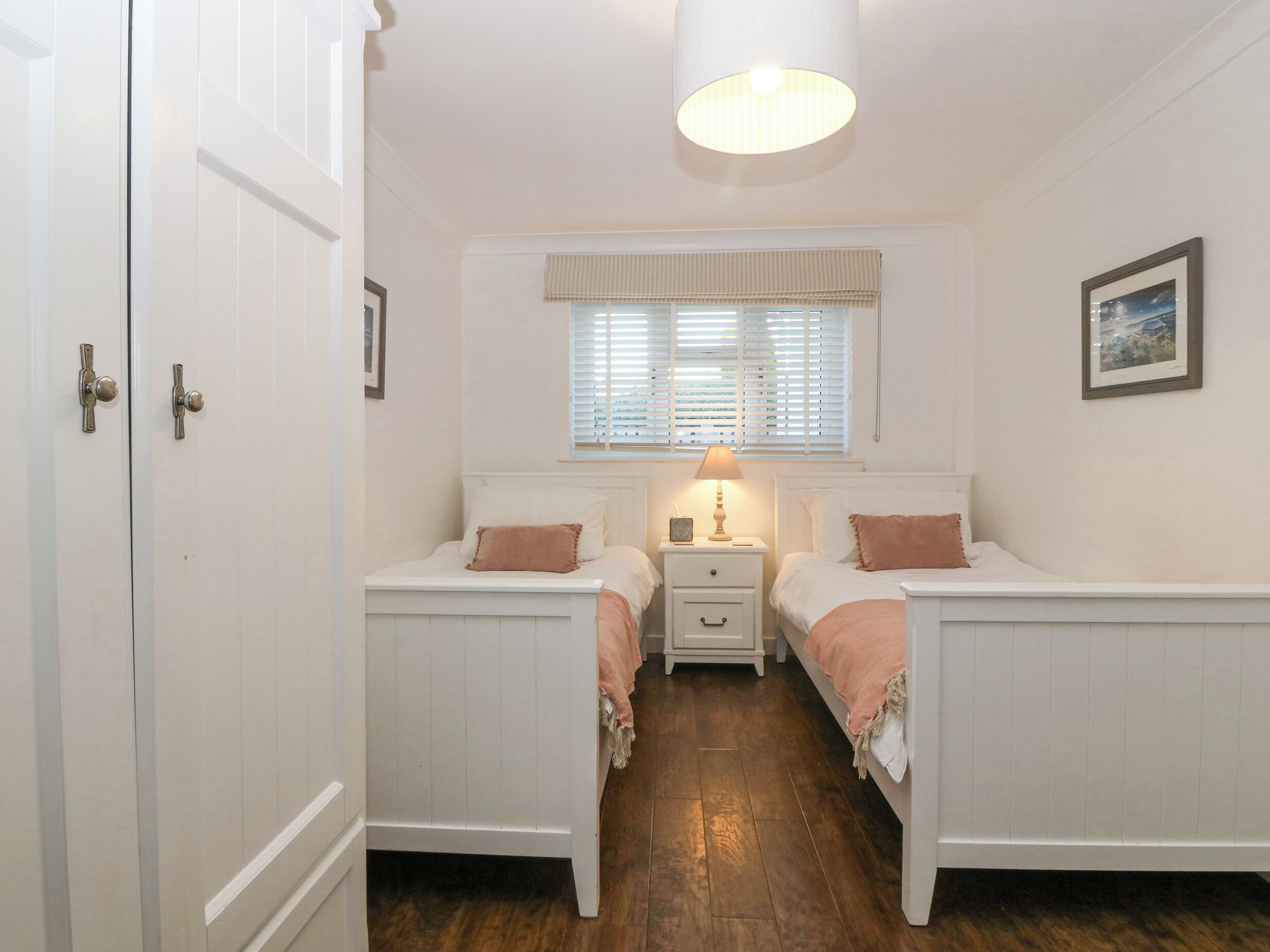 A bedroom with two single beds, a nightstand with a lamp, a window with blinds, and a framed picture on the wall.