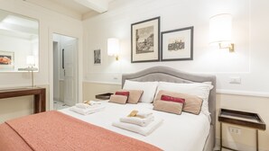 En pænt redt seng med hvide sengetøj og et pink pled, et natbord med lampe, og billeder i rammer på væggen.