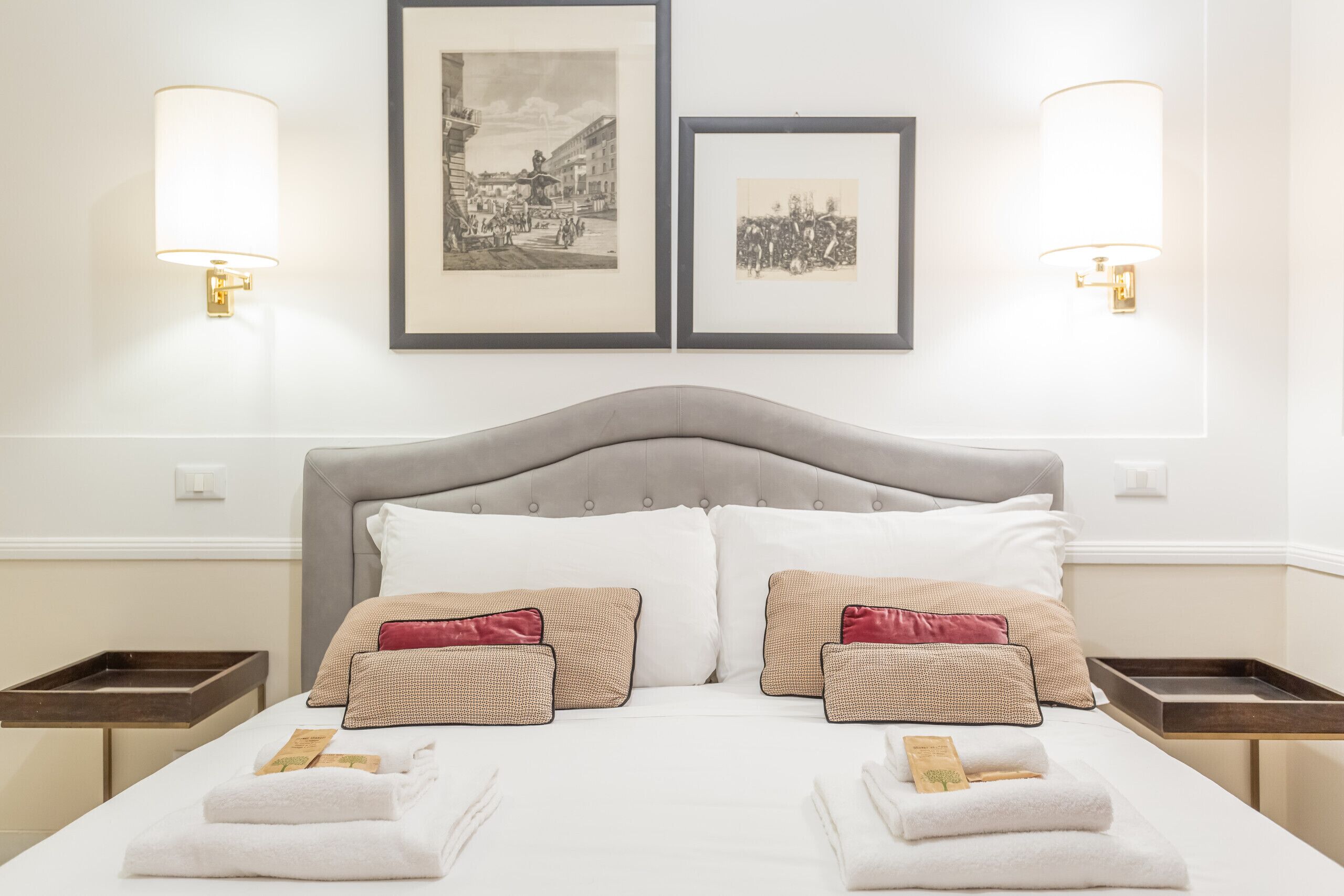 A neatly made bed with white linens and two decorative pillows, a framed black and white photo above it, and two wall-mounted lamps on either side.
