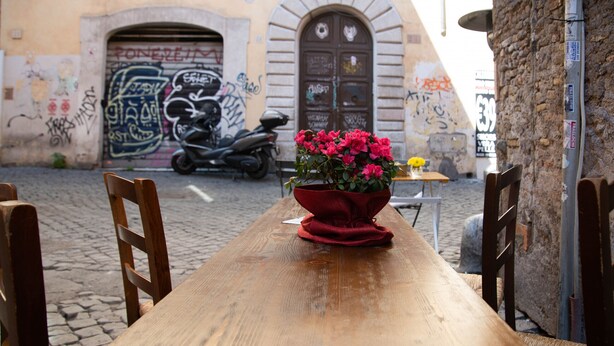Ein Holztisch mit Stühlen, ein roter Topf mit Blumen und ein Roller vor einem Gebäude mit Graffiti.