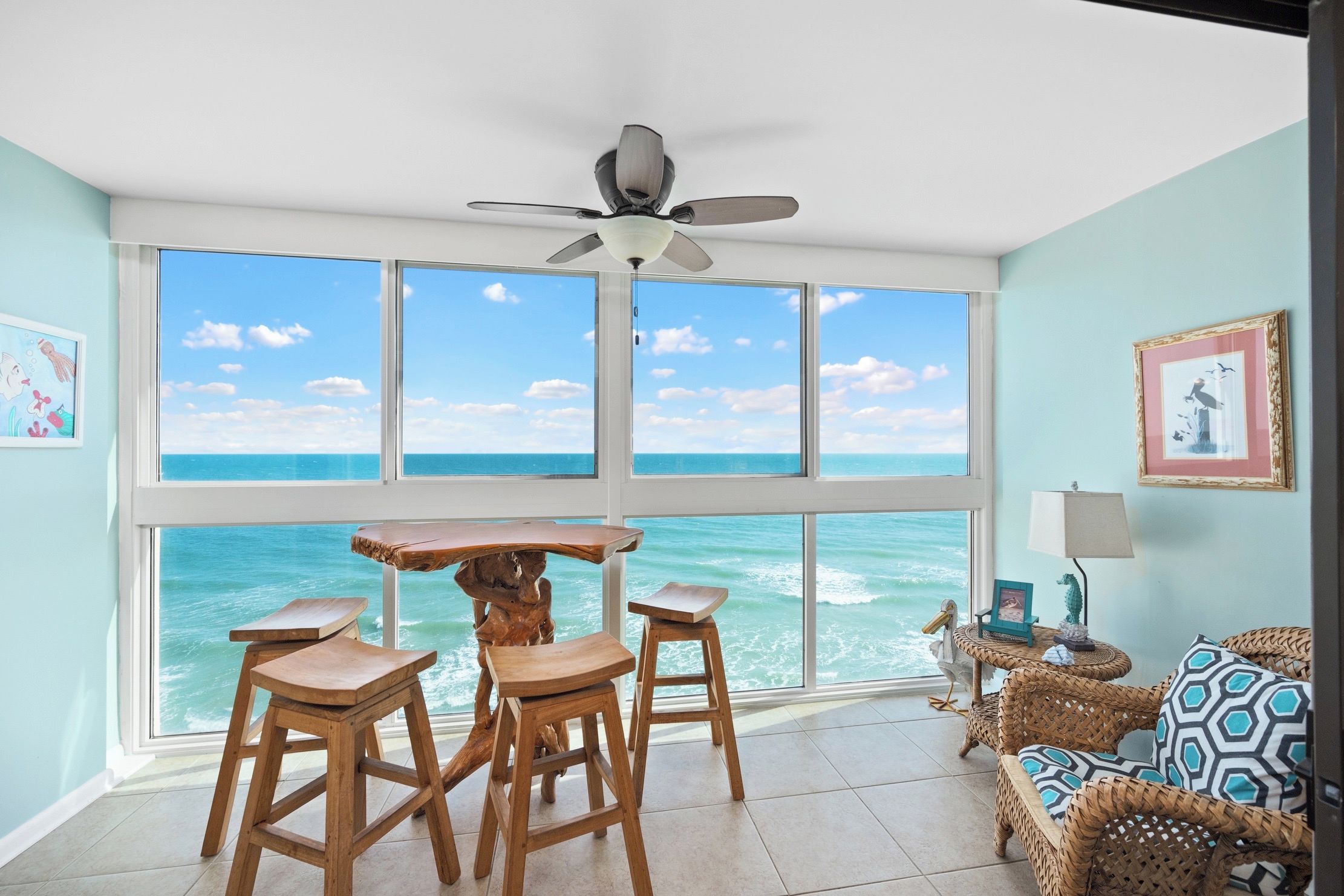 A room with a large window overlooking the ocean, wooden furniture, a ceiling fan, and framed artwork.