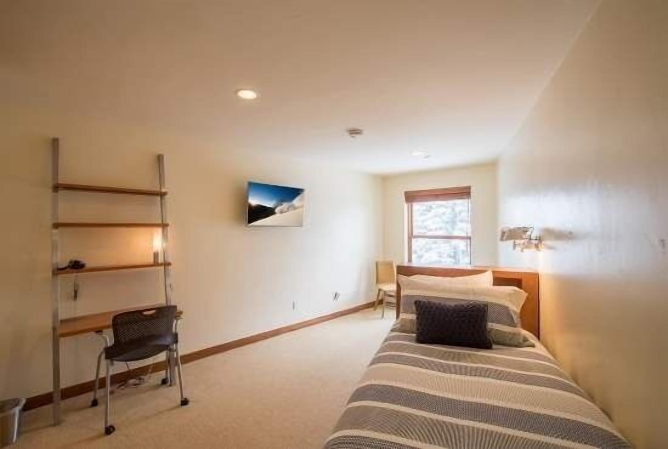 Un dormitorio con cama, escritorio, silla, ventana y un cuadro en la pared.