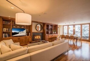 A spacious living room with a fireplace, bookshelves, a large TV, and a dining area with natural light from large windows.