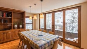 A dining area with a table set for four, a wooden cabinet with books, and a view of a snowy balcony. - Northwoods Private Condo with Easy Ski-In Ski-Out Access by RedAwning (Vail)