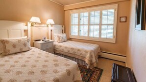 Un dormitorio con dos camas, una mesita de noche, una ventana con persianas y una alfombra pequeña.