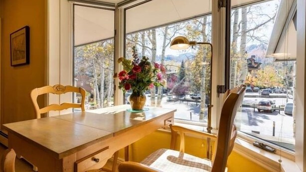 Un comedor con una mesa y sillas de madera, un jarrón con flores y vista a un estacionamiento a través de grandes ventanales.