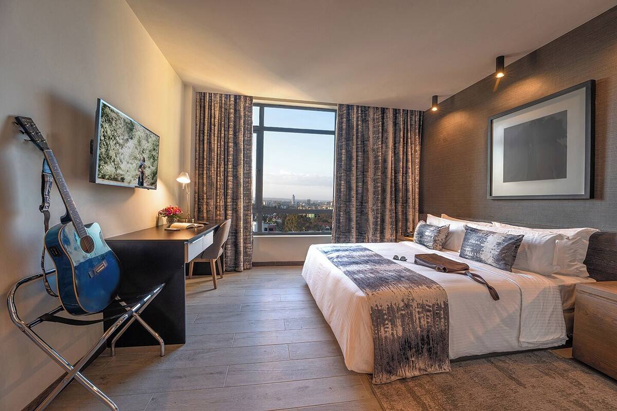 Ein Hotelzimmer mit Bett, Schreibtisch, Gitarre und Blick auf die Stadt durch das Fenster.
