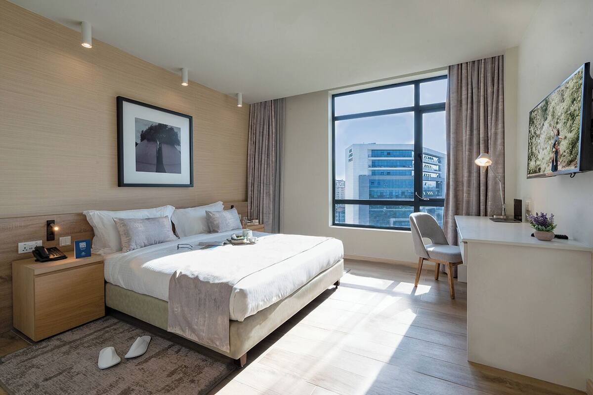 Ein modernes Hotelzimmer mit einem großen Bett, Nachttisch, Schreibtisch, Stuhl und einem großen Fenster mit Blick auf die Stadt.