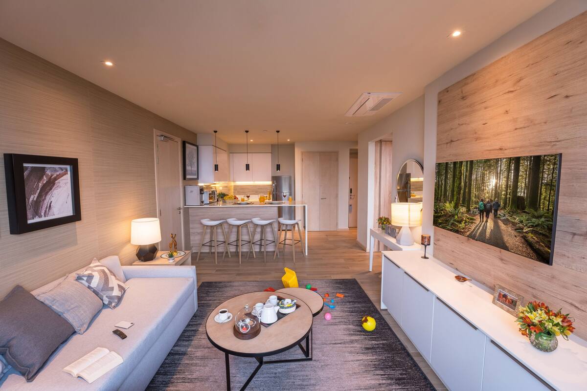 A modern hotel room with a sofa, a coffee table, a TV, and a kitchen area in the background.