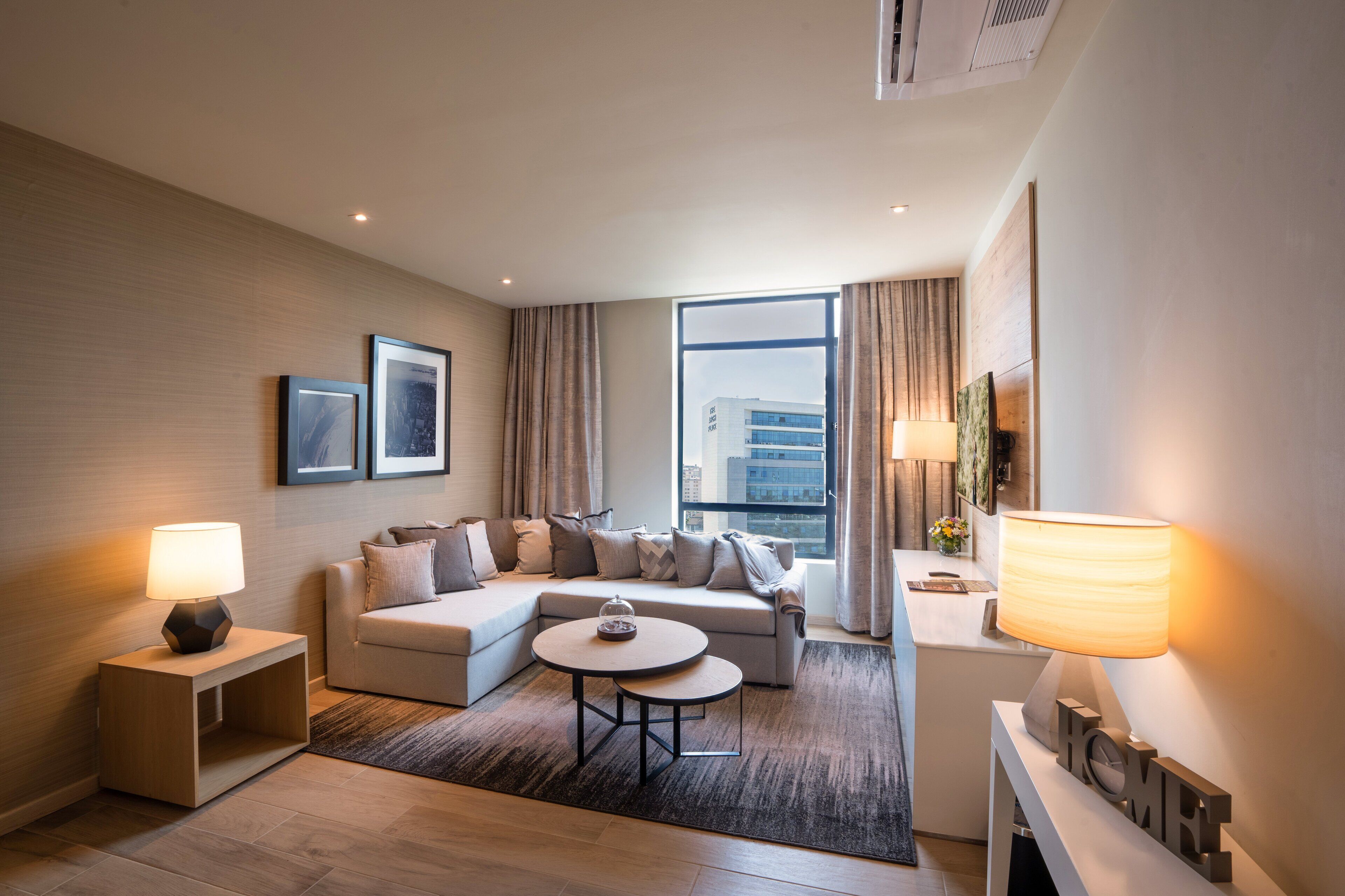 A modern hotel room with a sectional sofa, a round coffee table, a small side table with a lamp, and a large window offering a city view.