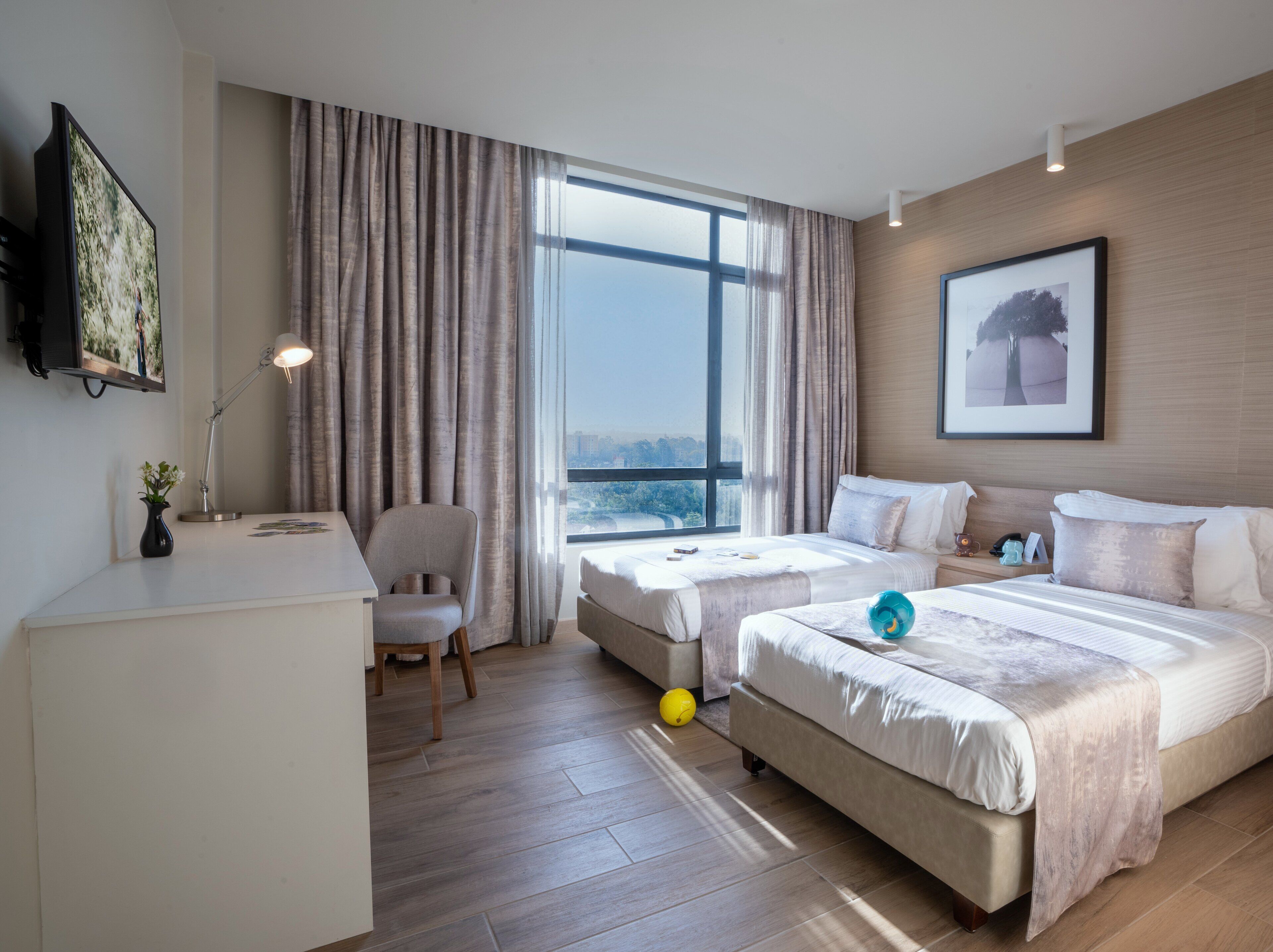 Ein Hotelzimmer mit zwei Betten, einem Schreibtisch, einem Stuhl, einem Fernseher und einem großen Fenster mit Vorhängen.