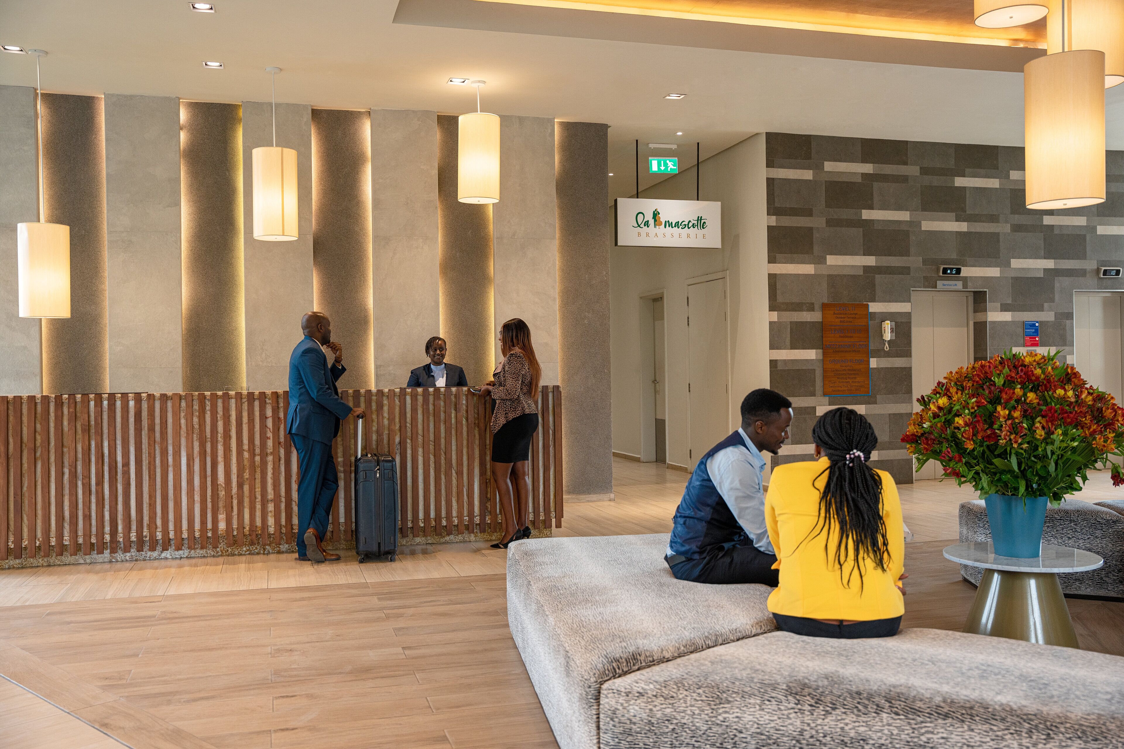 A hotel reception area with a wooden reception desk, a potted plant, and a seating area with a sofa and armchair.