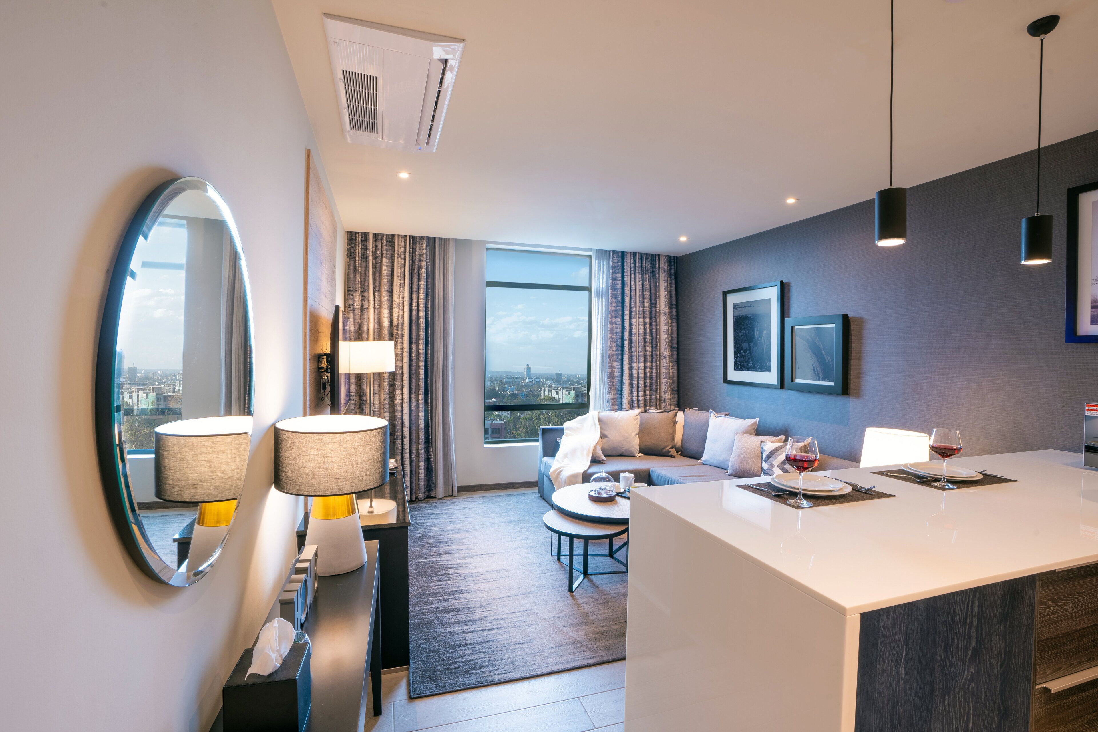 A modern hotel room with a large mirror, a sofa, a dining table, and a city view.