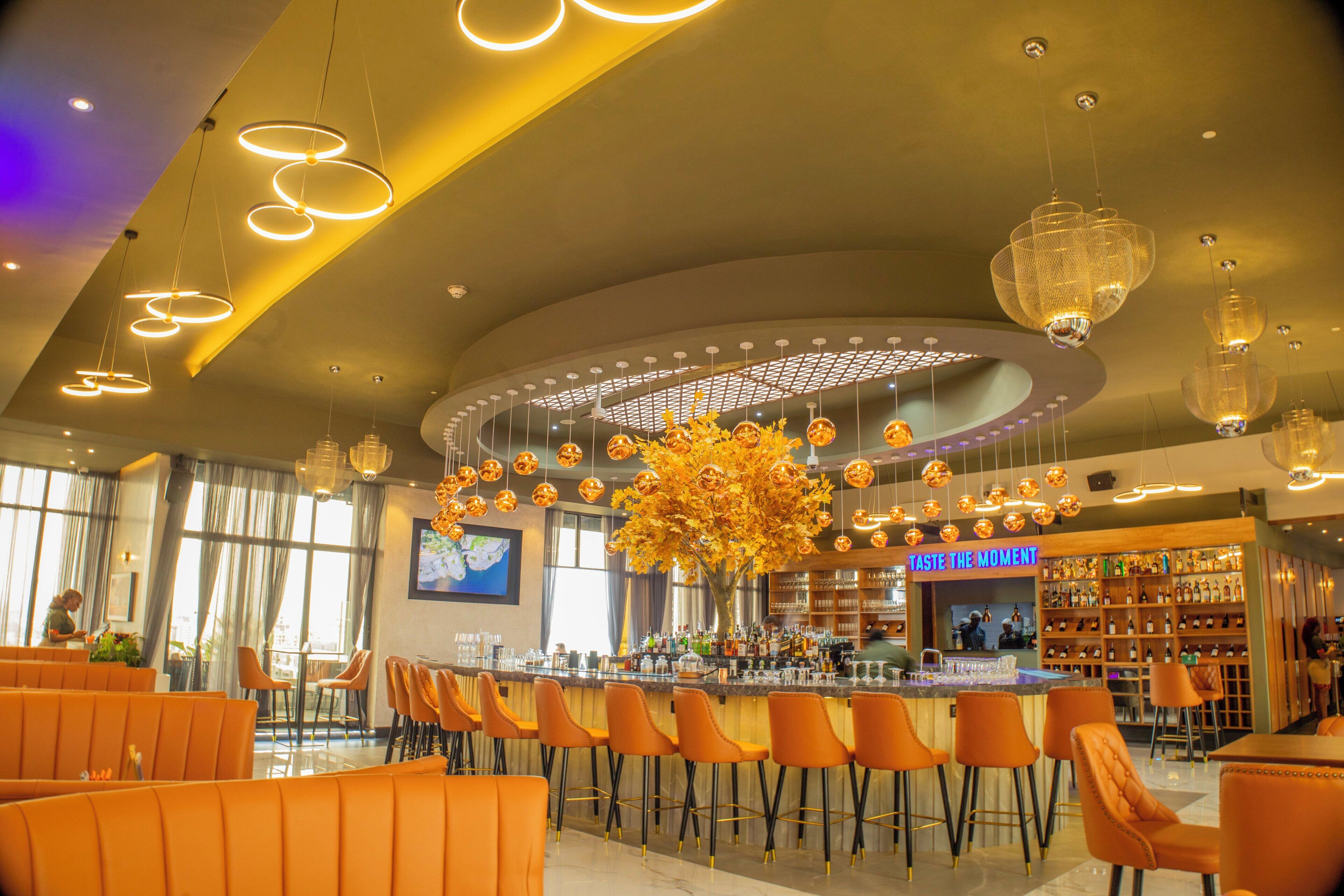 Zona de bar moderna con asientos naranja, una barra central y un arreglo de plantas decorativo.