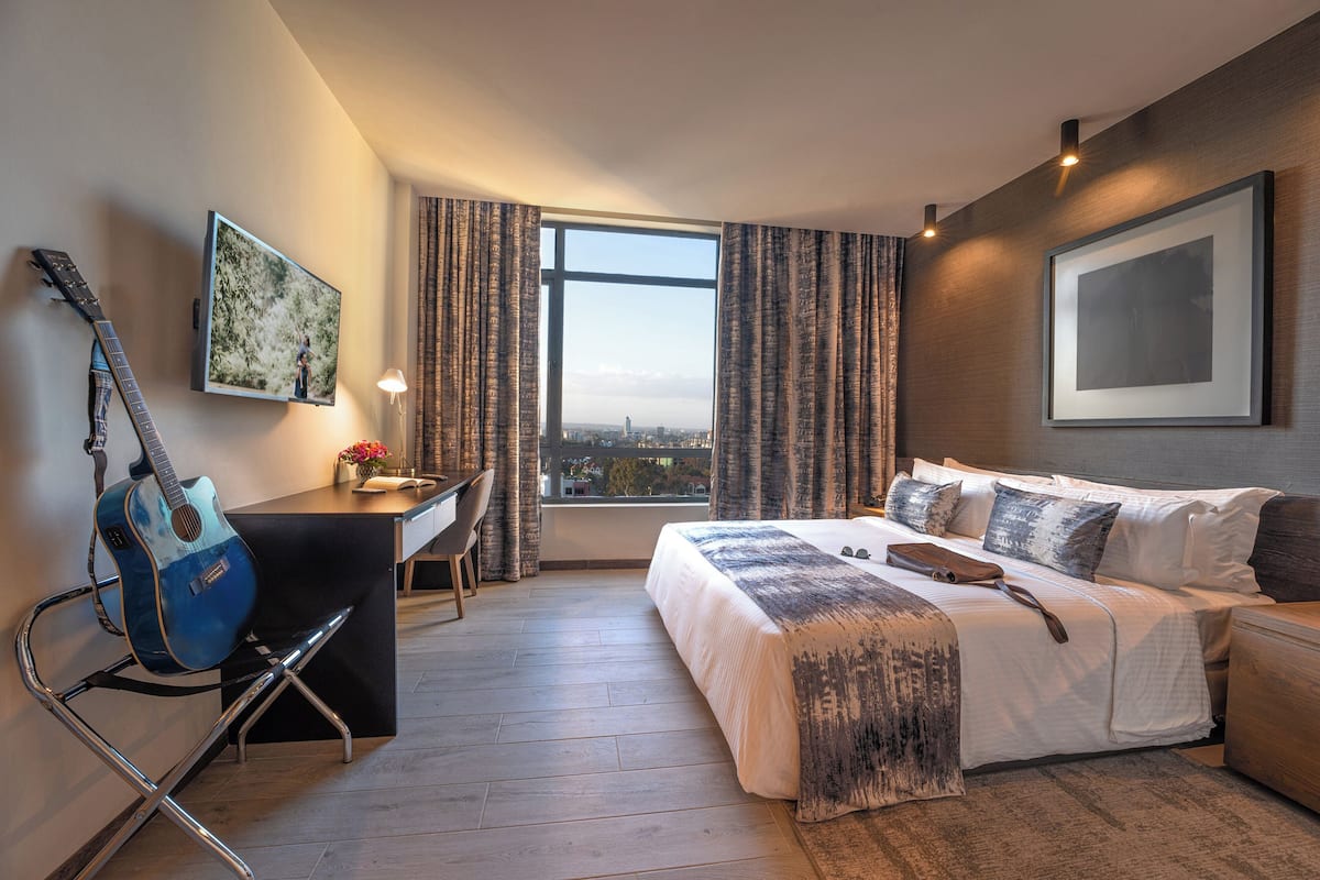A hotel room with a bed, a desk, a chair, a guitar, and a large window with curtains.