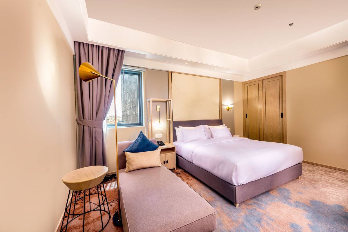 Ein modernes Hotelzimmer mit einem großen Bett, einem kleinen Schreibtisch, einer Bank und einem großen Fenster mit Vorhängen.