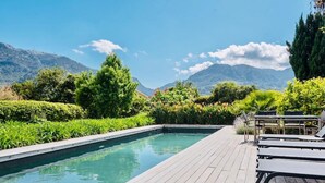 A pool with a wooden deck and lounge chairs, surrounded by lush greenery and mountains in the background.
