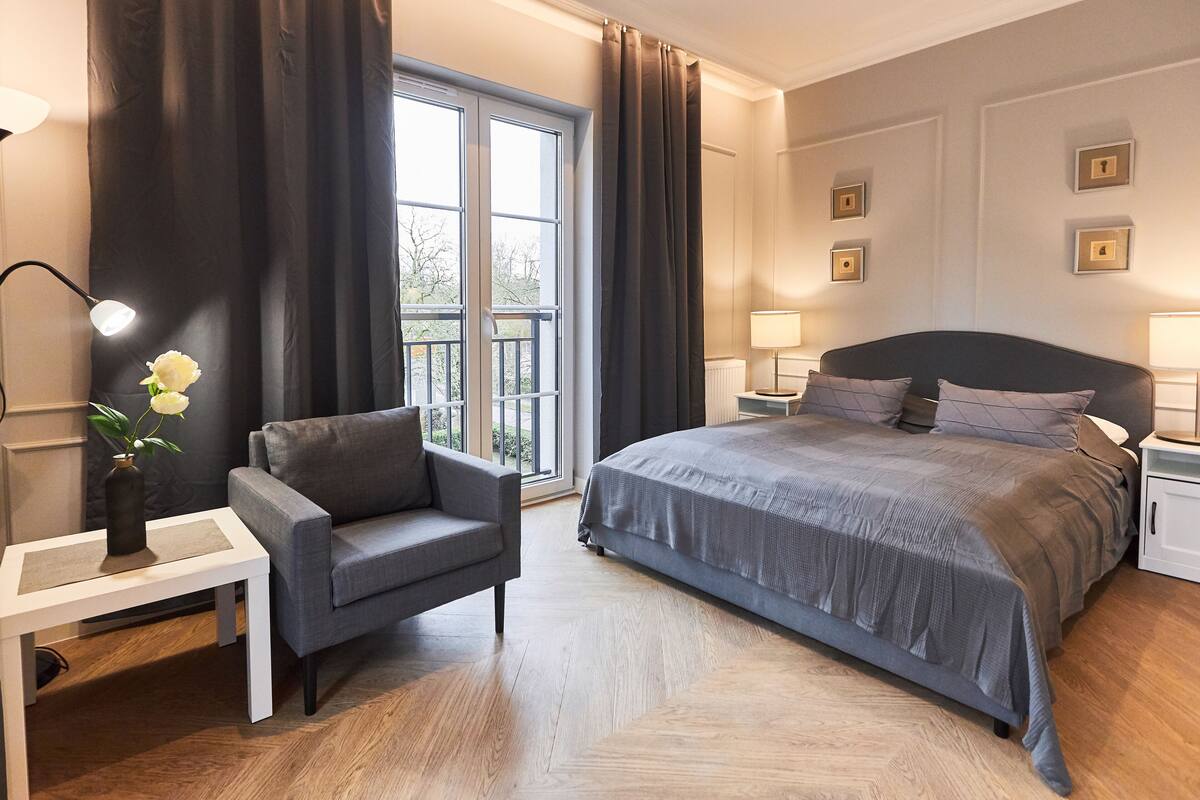 A modern bedroom with a large bed, a grey armchair, a white bedside table, and a window with curtains.