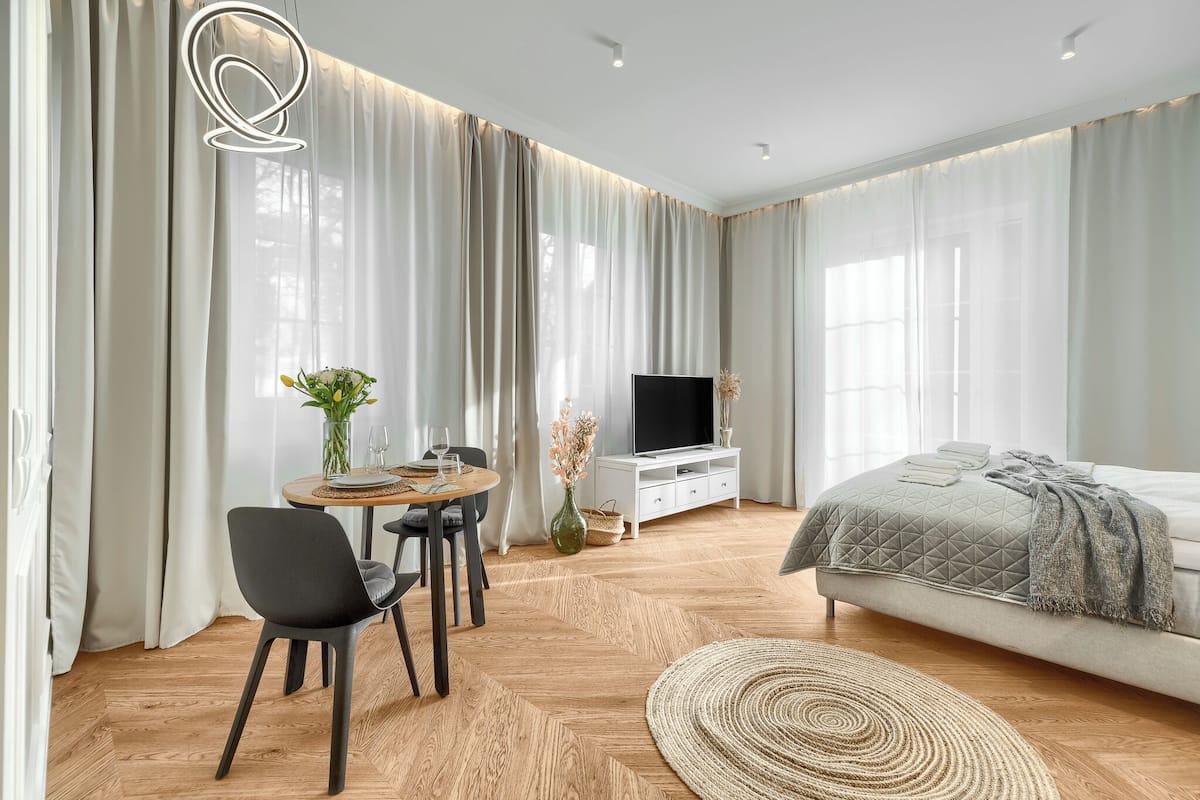 A modern bedroom with a bed, a TV, a dining area with chairs and a table, and a large window with curtains.