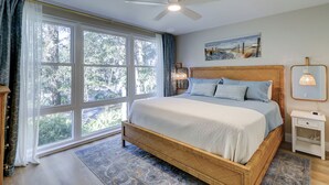Ein Schlafzimmer mit großem Fenster, Deckenventilator, Bett mit Holz-Kopfschild, Nachttisch mit Lampe und einem Bild an der Wand.