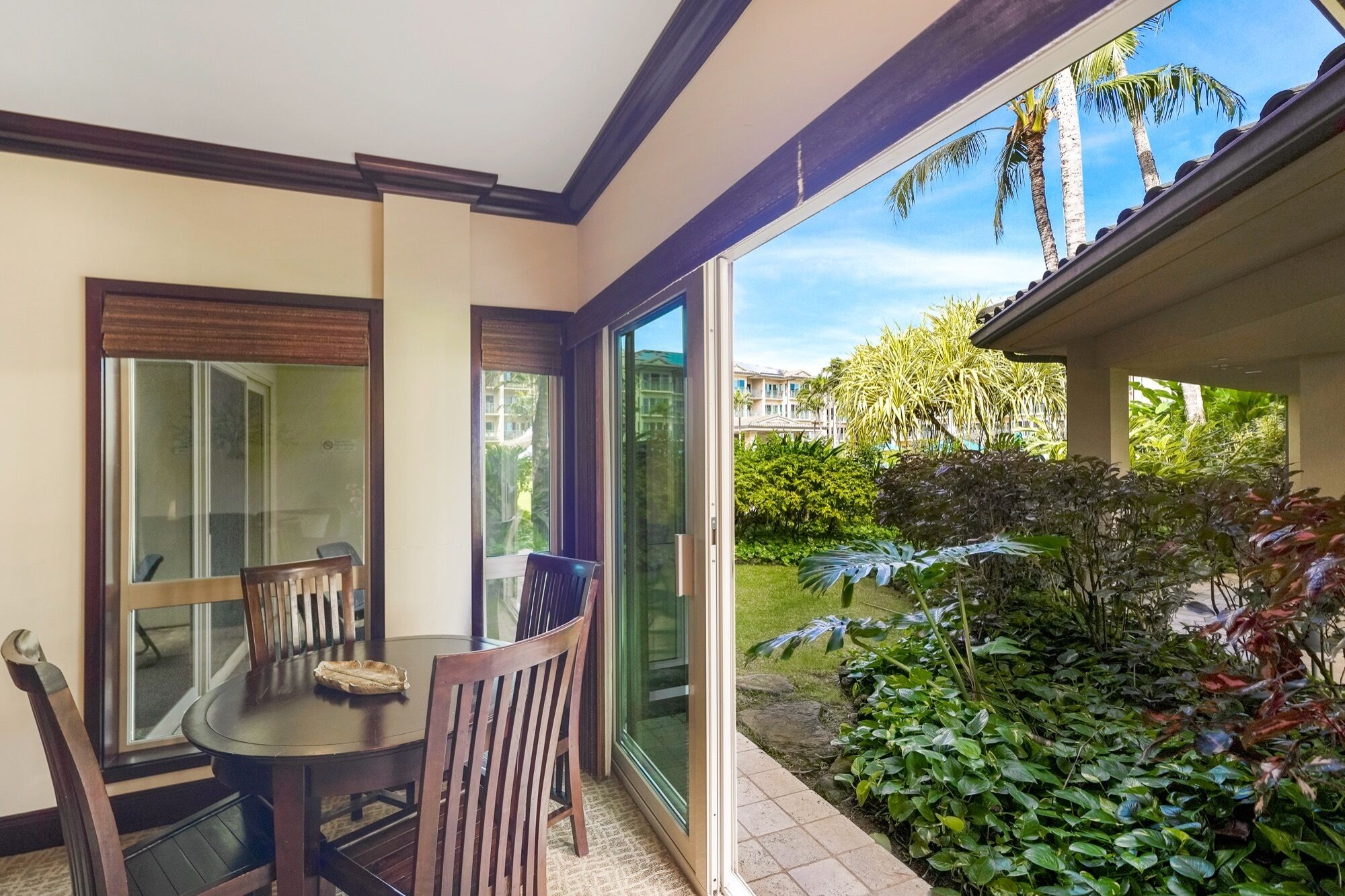 A dining area with a table and chairs, a sliding glass door leading to a garden with tropical plants and a view of a residential area.
